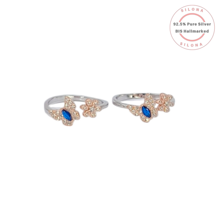 A pair of silver toe rings with a rose-gold plated butterfly and flower motif, featuring a central marquise-cut blue stone, shown on a white background with the Silona logo.