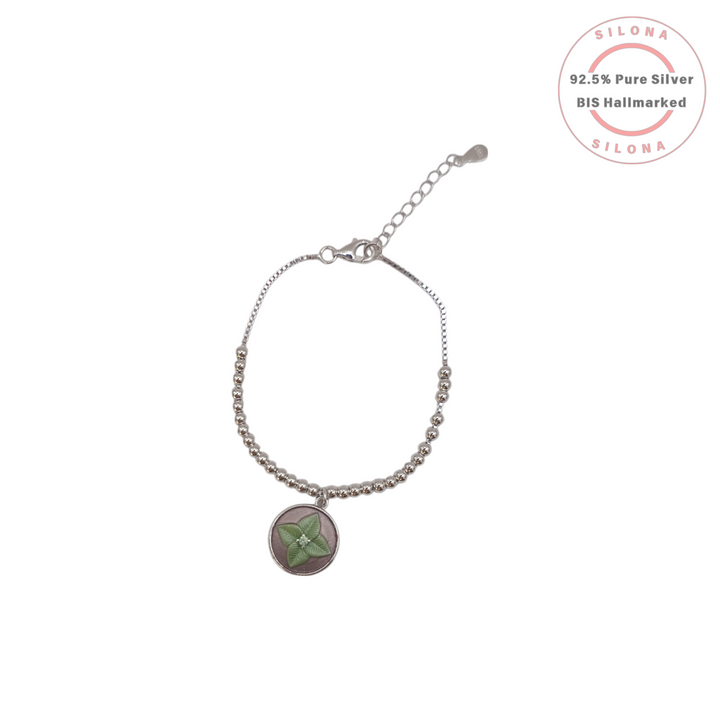 A beaded silver bracelet with an adjustable clasp featuring a round charm with a green four-leaf clover on a mauve-pink background.