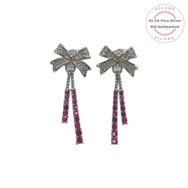 A pair of silver bow dangle earrings, with the bows studded in cubic zirconia and tassels of ruby-hued stones, on a white background.