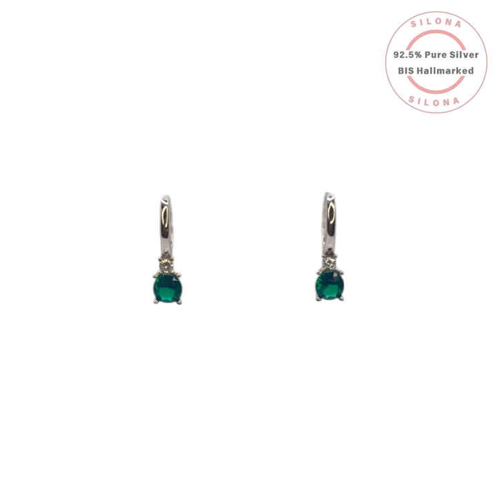 A pair of Silona silver huggie hoop earrings, each with a dangling oval-cut emerald green gemstone and a small clear crystal accent, on a white background.