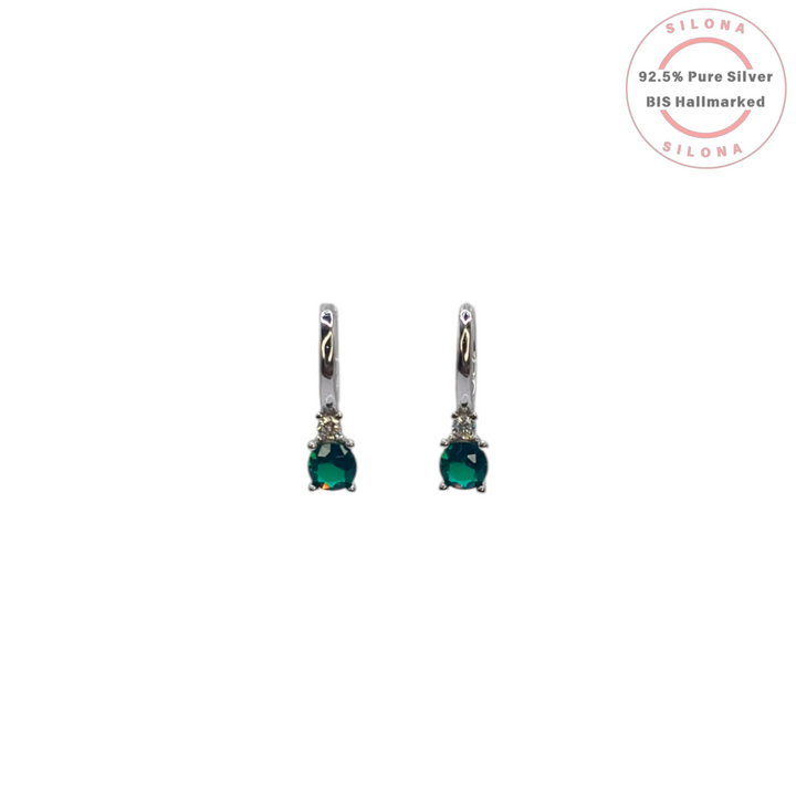 Pair of Silona silver huggie hoop earrings with a drop featuring a round emerald-green stone and a small clear crystal, on a white background.