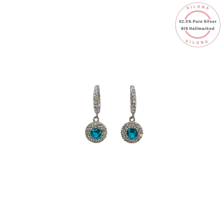 A pair of Silona silver huggie hoop earrings with a dangling round halo charm featuring a central aquamarine blue cubic zirconia stone, on a white background.
