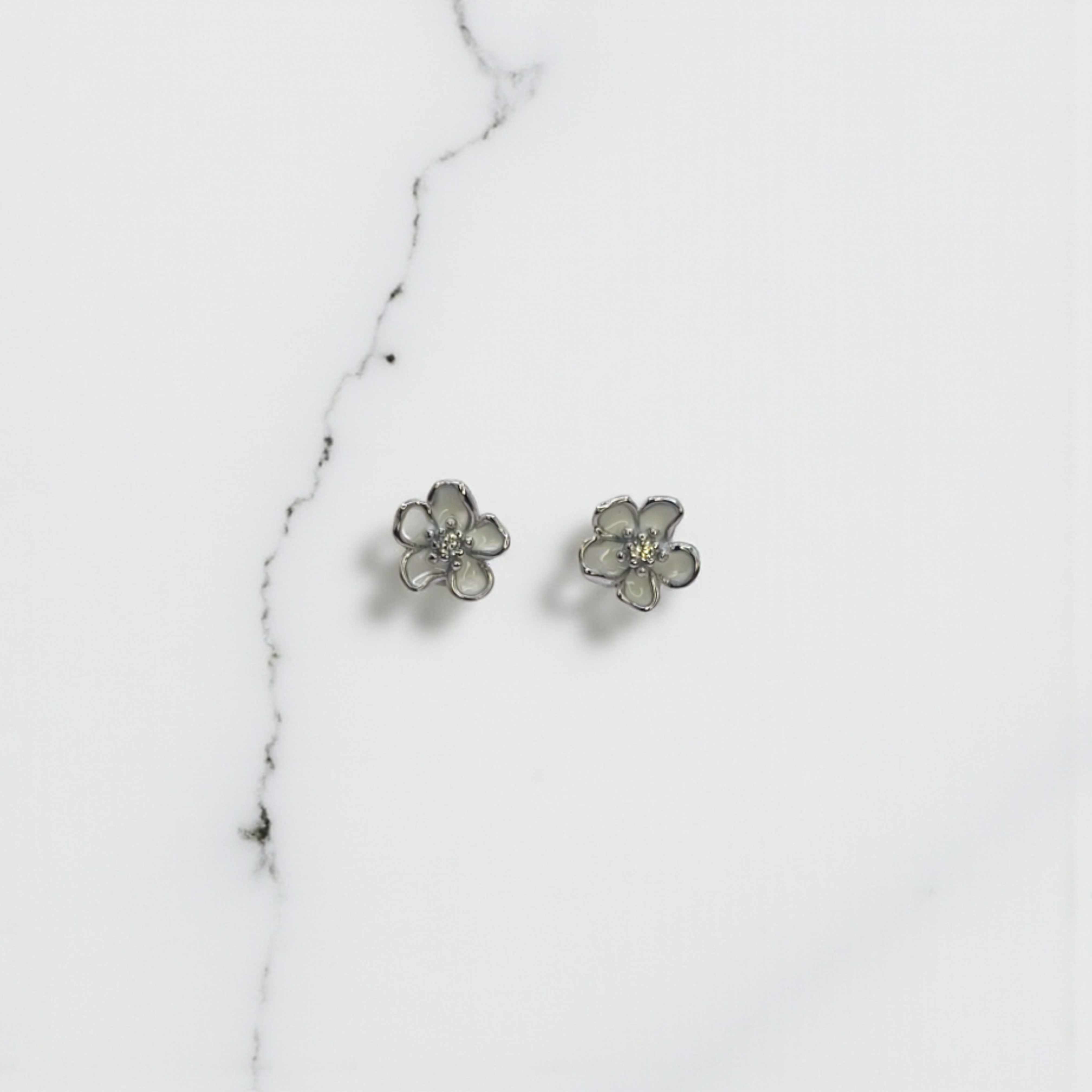 A pair of Ethereal Blossom Stud Earrings in 92.5 silver, featuring a five-petal flower design with creamy enamel and a sparkling center, displayed on a white marble surface.