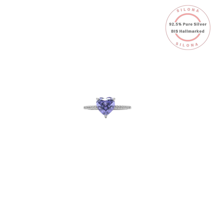 A silver solitaire ring featuring a large, heart-shaped, violet-blue tanzanite-hued stone on a pave-set cubic zirconia band.