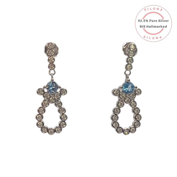 A pair of Silona silver drop earrings featuring a linear setting of white cubic zirconia, leading to an open loop motif set with more white CZ and a central light blue cubic zirconia stone, on a white background.